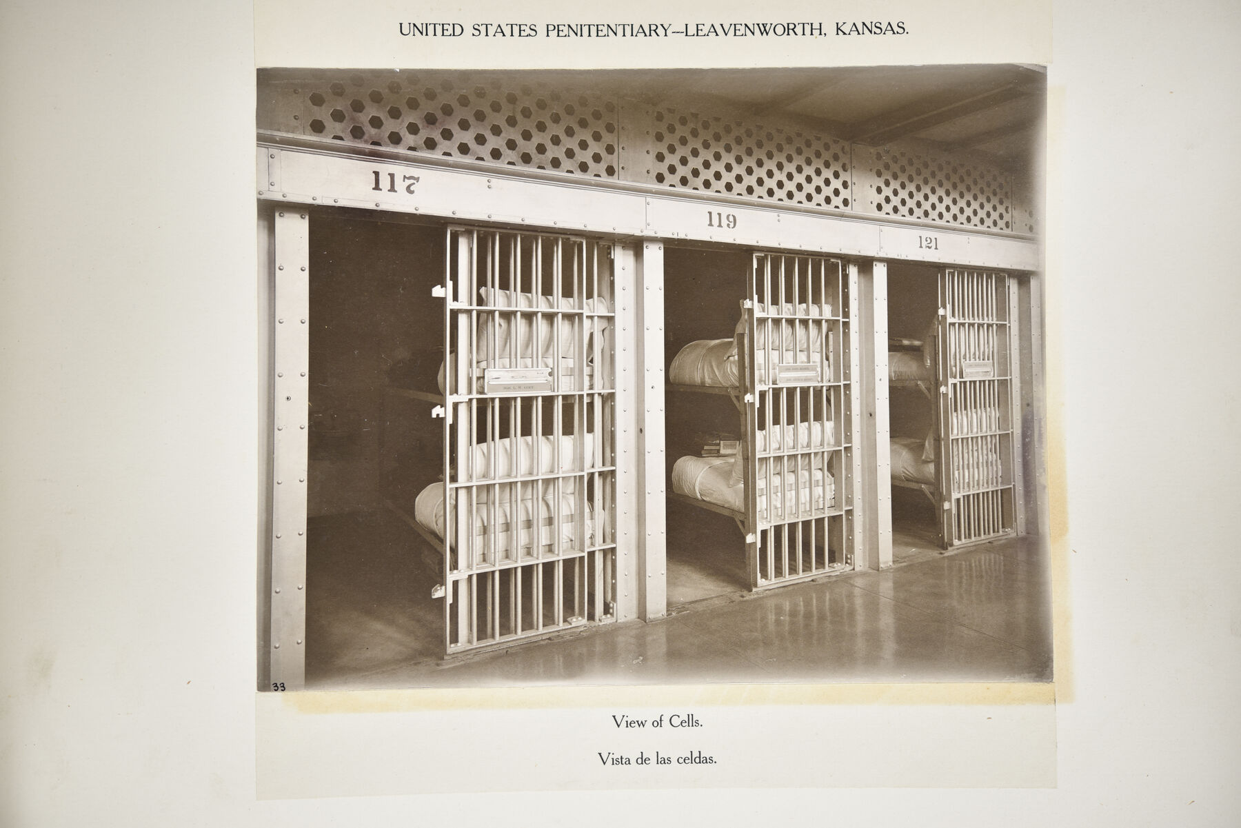 Mounted photograph of three open cells at Leavenworth with numbers 117, 119 and 121 above them and folded bedding on the bunks.