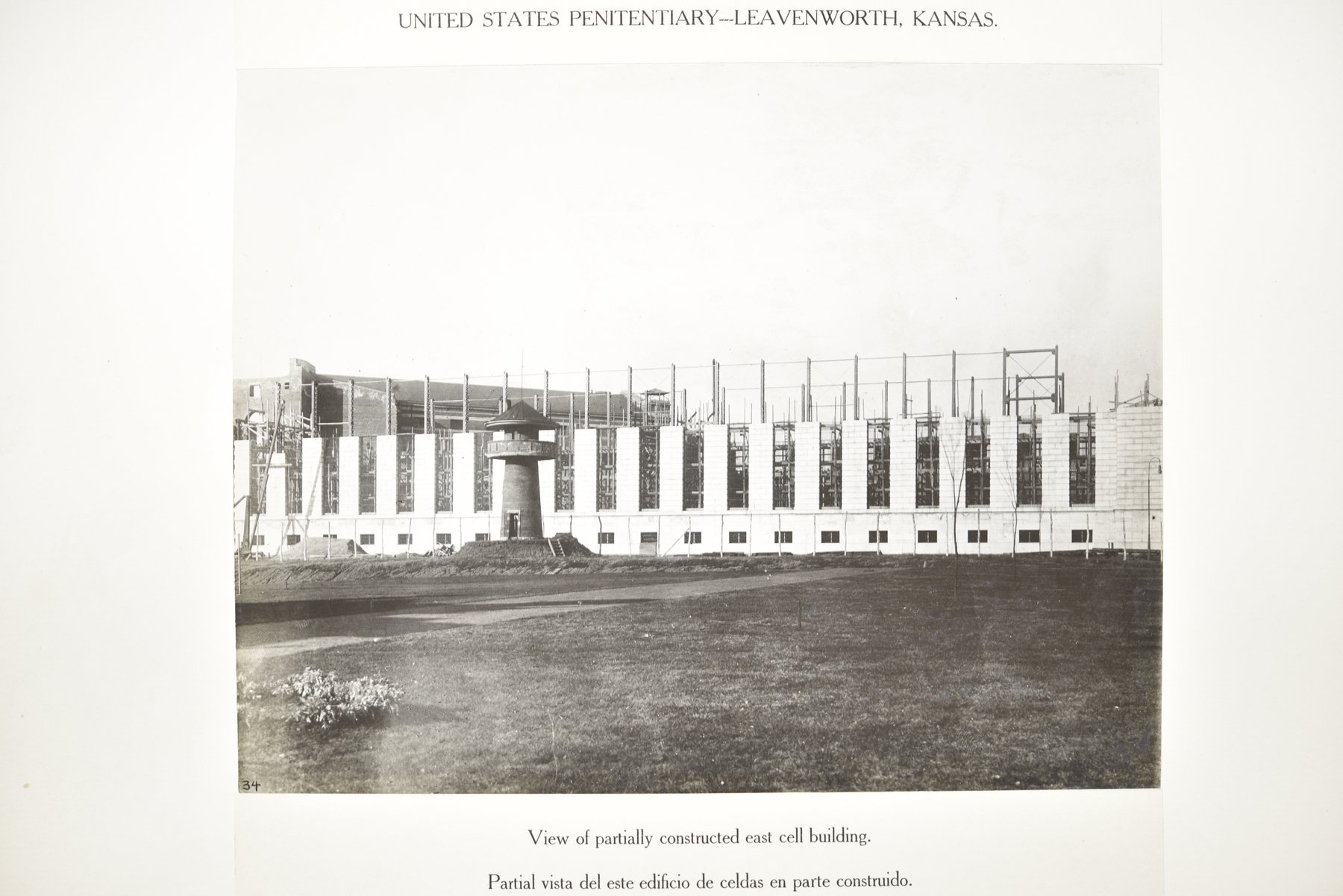 Album page headed United States Penitentiary, Leavenworth, Kansas, with a photograph of a long building with stone piers and open steel framing above, a round guard tower with a conical roof in front of it, and a lawn and shrub in the foreground, above the caption View of partially constructed east cell building.
