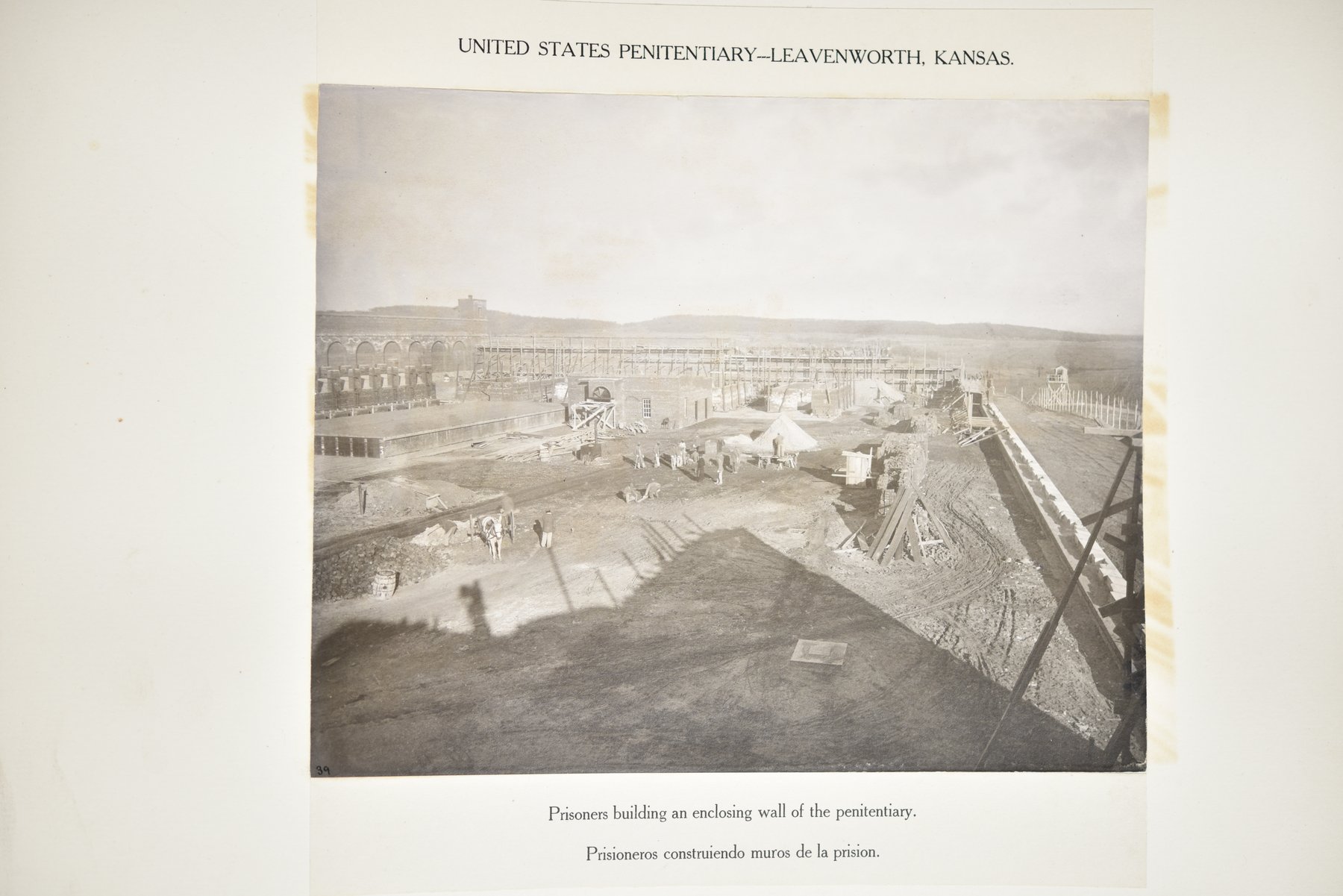 Album page headed United States Penitentiary, Leavenworth, Kansas, with a photograph of a high view over a construction site: a long wall under scaffolding, cell blocks with arched openings at the left, piles of sand and stone, and small figures of men working with carts and a horse, above the caption Prisoners building an enclosing wall of the penitentiary.