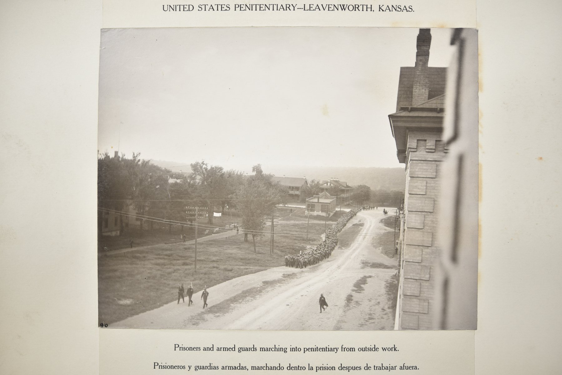 Album page headed United States Penitentiary, Leavenworth, Kansas, with a photograph of an elevated view from a stone building down a dirt road: a long column of prisoners marches toward the building, with figures along the road and in the field, and trees and houses beyond, above the caption Prisoners and armed guards marching into penitentiary from outside work.