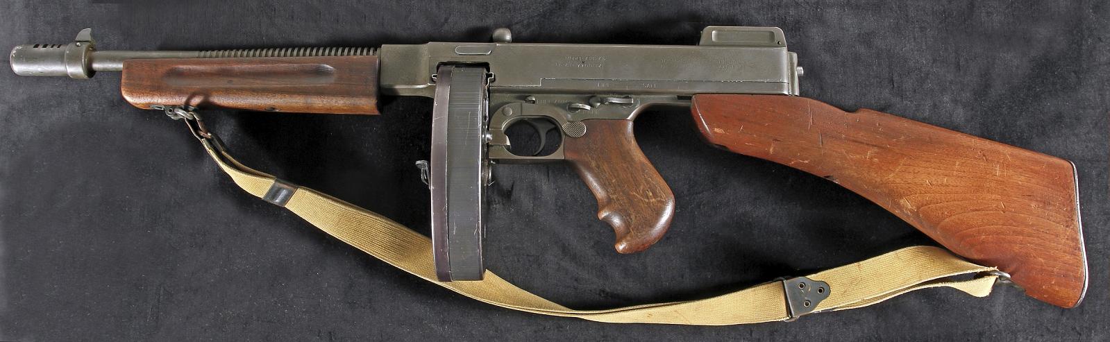 A Thompson submachine gun with a drum magazine and a canvas strap, lying on dark cloth.